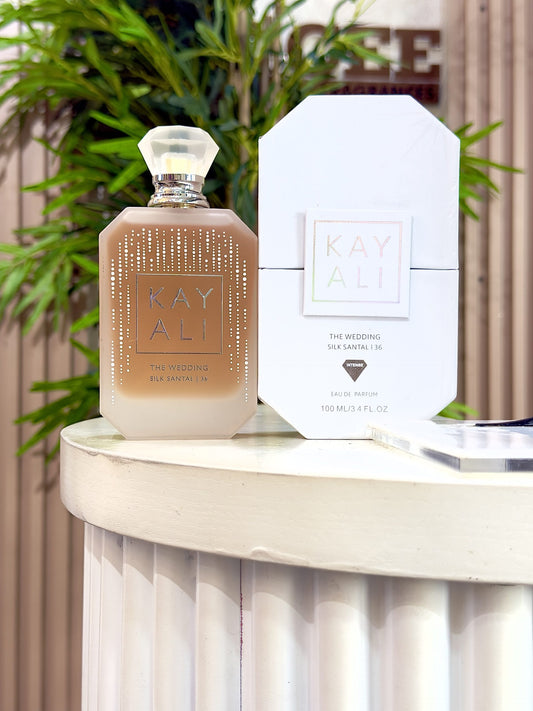 A bottle of Kay Ali The Wedding Eau de Parfum next to its white packaging box, displayed on a white pedestal with a blurred background of green foliage and a wooden wall.