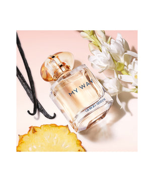 A perfume bottle labeled 'MY WAY' and 'GABRIELLE MYSTIQUE' placed on a pale pink background with white flowers, vanilla beans, and a pineapple slice around it.