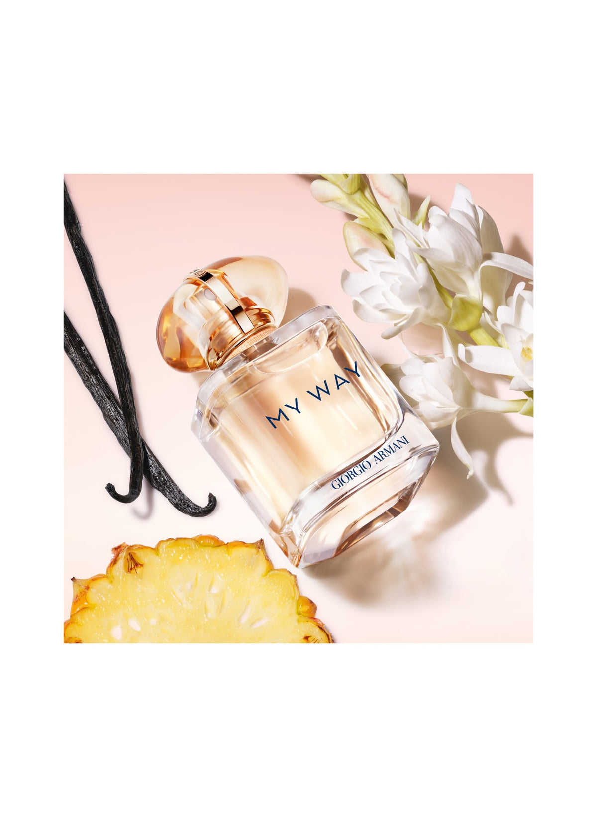 A perfume bottle labeled 'MY WAY' and 'GABRIELLE MYSTIQUE' placed on a pale pink background with white flowers, vanilla beans, and a pineapple slice around it.