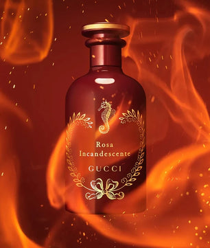 A Gucci Rosa Incandescente perfume bottle with a metallic rose-gold finish, featuring a seahorse emblem and gold laurel wreath detailing, set against a dramatic background of orange and red flames.
