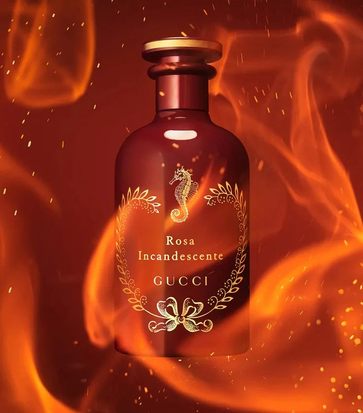 A Gucci Rosa Incandescente perfume bottle with a metallic rose-gold finish, featuring a seahorse emblem and gold laurel wreath detailing, set against a dramatic background of orange and red flames.
