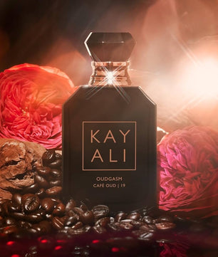 Perfume bottle labeled 'KAY ALI' surrounded by coffee beans and flowers on a dark background