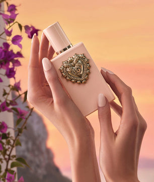Hand holding a pink perfume bottle with decorative cap against a blurred floral background