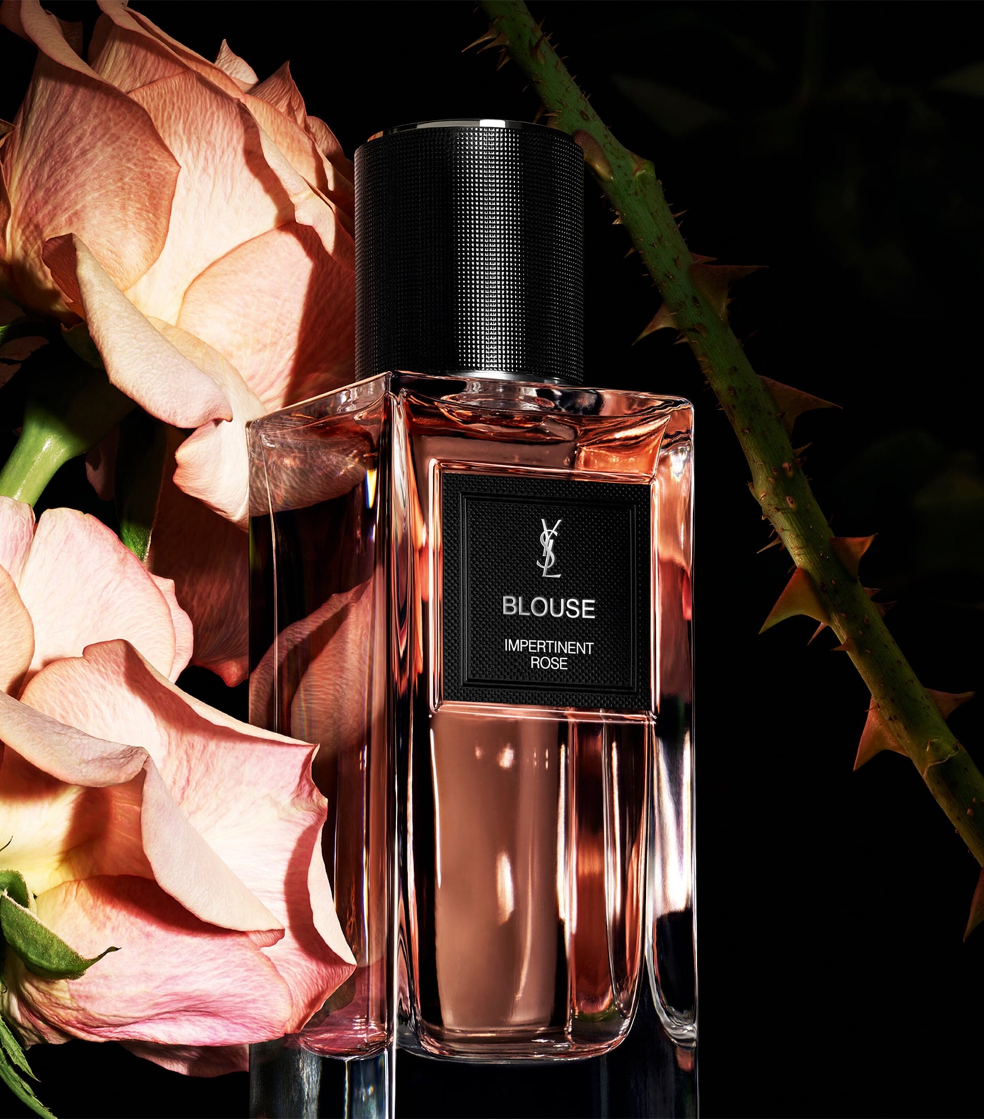 Perfume bottle with 'Yves Saint Laurent Blouse' label surrounded by pink roses on a dark background