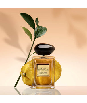 A bottle of Armani Privé Orange Mediterraneo perfume, styled with a fresh green citrus branch and two yellow oranges on a reflective surface against a warm beige background.