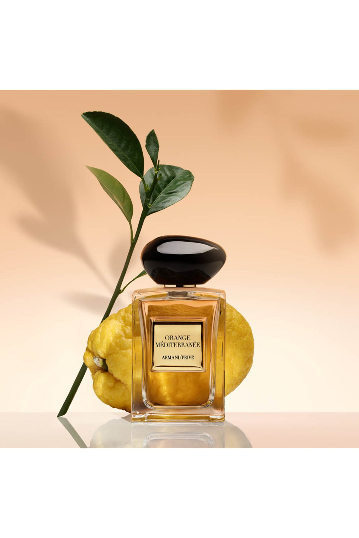 A bottle of Armani Privé Orange Mediterraneo perfume, styled with a fresh green citrus branch and two yellow oranges on a reflective surface against a warm beige background.