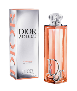 Dior Addict Peachy Glow Eau de Parfum bottle and packaging box on a white background. The rectangular bottle is filled with peach-colored liquid and features a silver Dior logo, while the box is metallic silver with peach accents and black text.