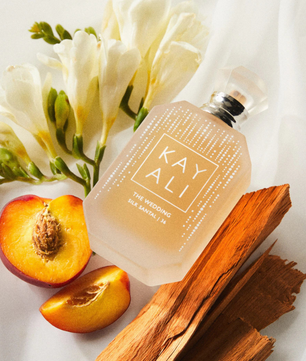 A frosted glass perfume bottle labeled 'KAY ALI THE WEDDING SILK BATTLE IN' arranged on a white fabric background with white flowers, sliced peach, and wood pieces.