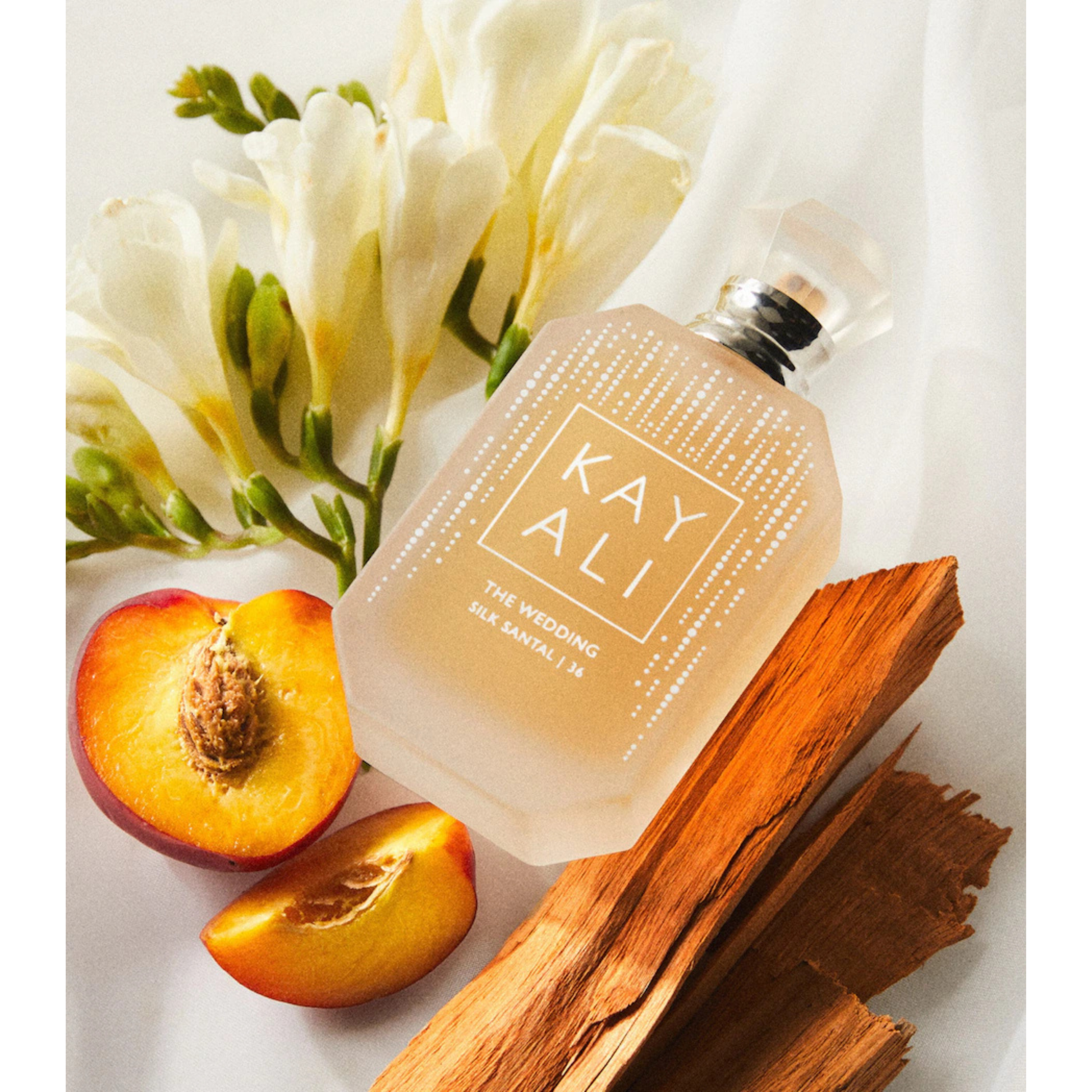 A frosted glass perfume bottle labeled 'KAY ALI THE WEDDING SILK BATTLE IN' arranged on a white fabric background with white flowers, sliced peach, and wood pieces.