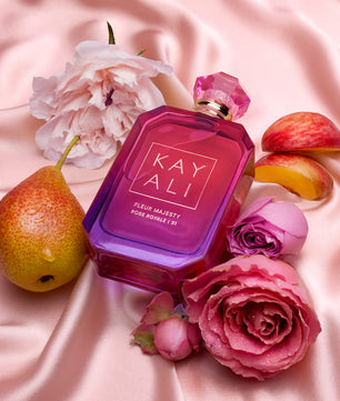 Perfume bottle labeled 'KAY ALI' surrounded by flowers and fruits on a pink fabric background