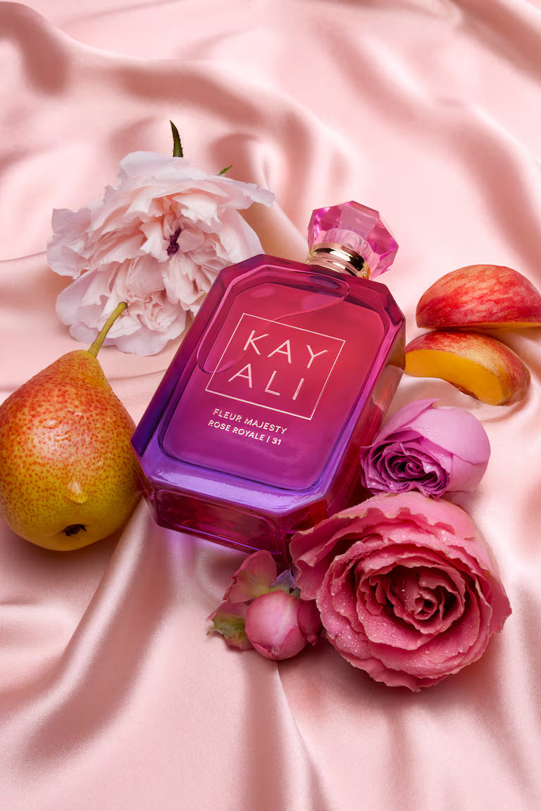 Perfume bottle labeled 'KAY ALI' surrounded by flowers and fruits on a pink fabric background