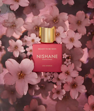 Nishane 'Meaning to Be Seen' Eau de Parfum bottle, featuring a vibrant pink rectangular body and a metallic gold cap, nestled among soft pink cherry blossoms.
