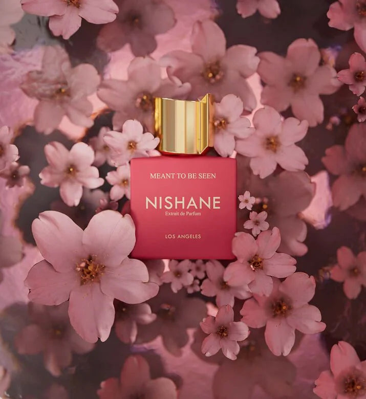 Nishane 'Meaning to Be Seen' Eau de Parfum bottle, featuring a vibrant pink rectangular body and a metallic gold cap, nestled among soft pink cherry blossoms.