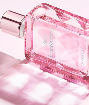 A Givenchy Very Irresistible perfume bottle in a faceted pink glass design with a metallic rose gold cap, set against a soft pink background with a reflective surface.