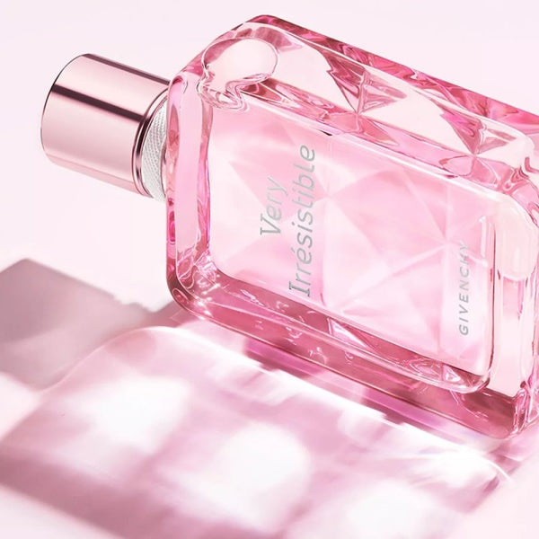 A Givenchy Very Irresistible perfume bottle in a faceted pink glass design with a metallic rose gold cap, set against a soft pink background with a reflective surface.