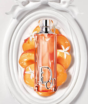 Dior Eau de Parfum bottle in a vibrant peach-orange color with a silver-tone Dior logo, presented on a white decorative background with peach slices.