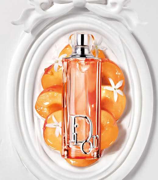 Dior Eau de Parfum bottle in a vibrant peach-orange color with a silver-tone Dior logo, presented on a white decorative background with peach slices.
