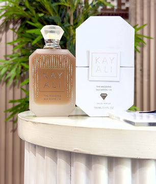 A bottle of Kay Ali The Wedding Eau de Parfum next to its white packaging box, displayed on a white pedestal with a blurred background of green foliage and a wooden wall.